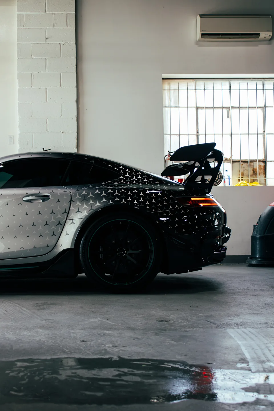A camouflage-wrapped Mercedes-AMG Black Series inside a lit garage, rear wing raised.