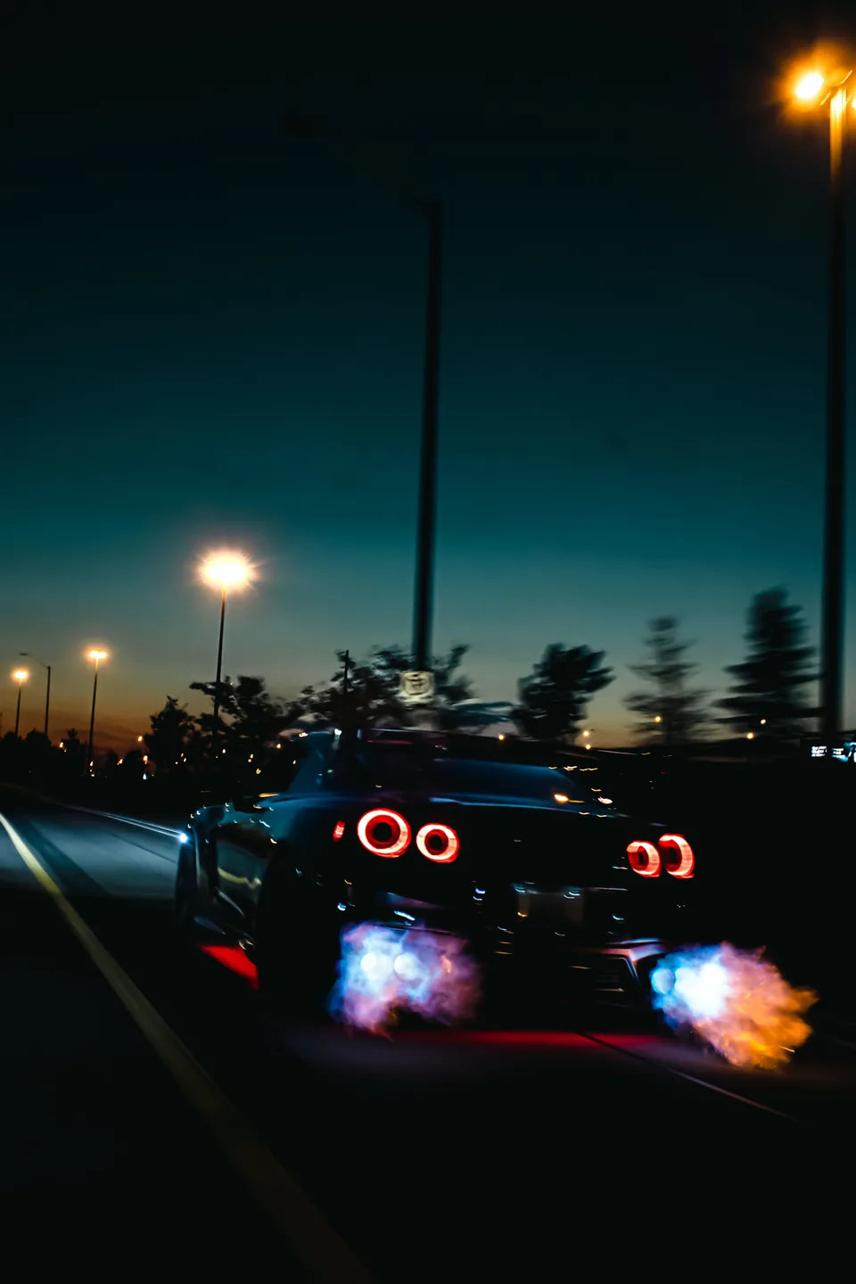 A Nissan GT-R shot from behind at night, flames from both exhausts and tail lights streaking as it pulls away.