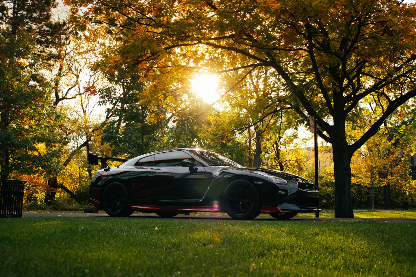 A Nissan GT-R parked on grass under autumn trees with the low sun burning through behind it.