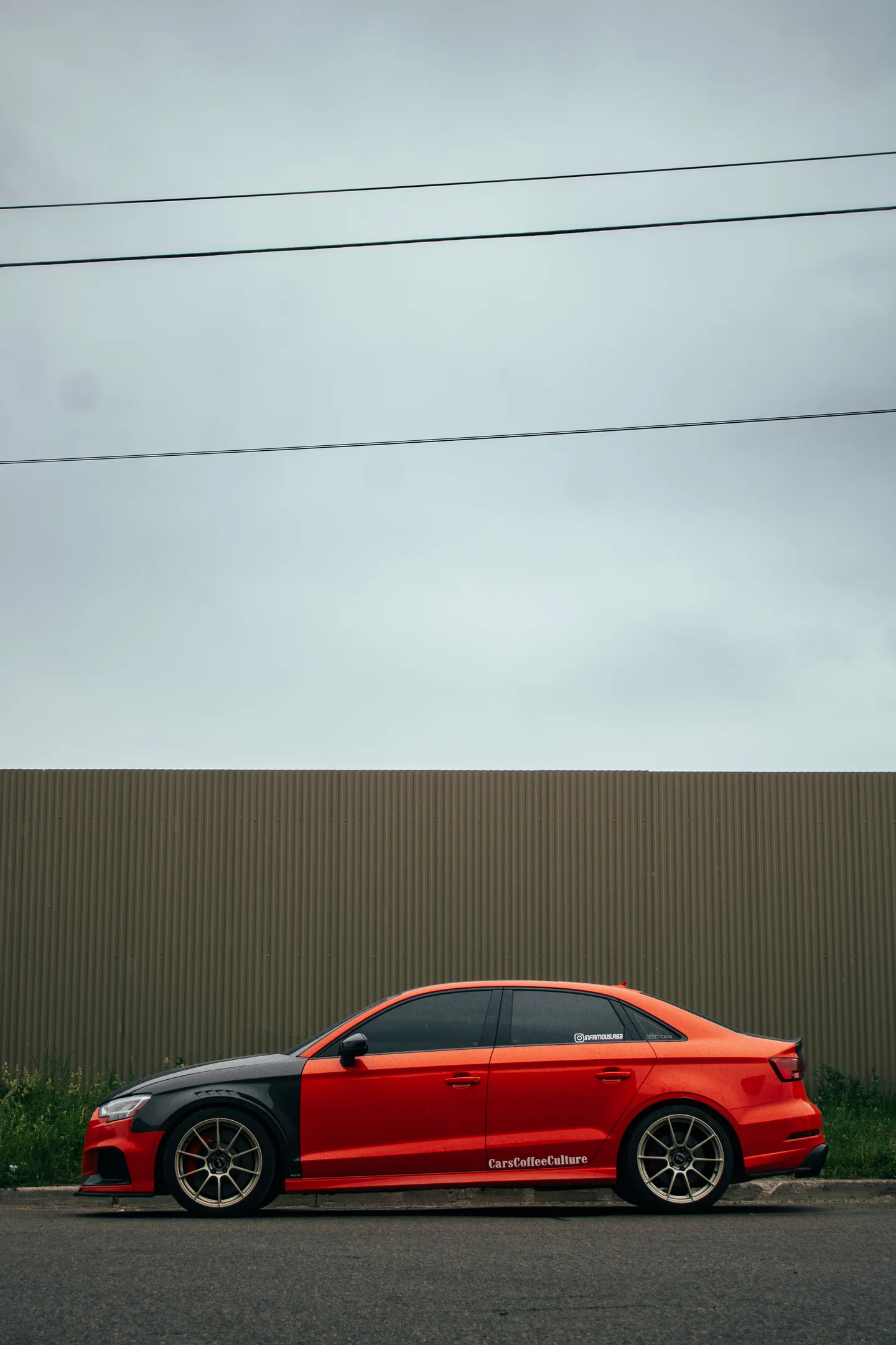 A red Audi RS3 in profile against a grey industrial wall with overhead lines crossing the sky.