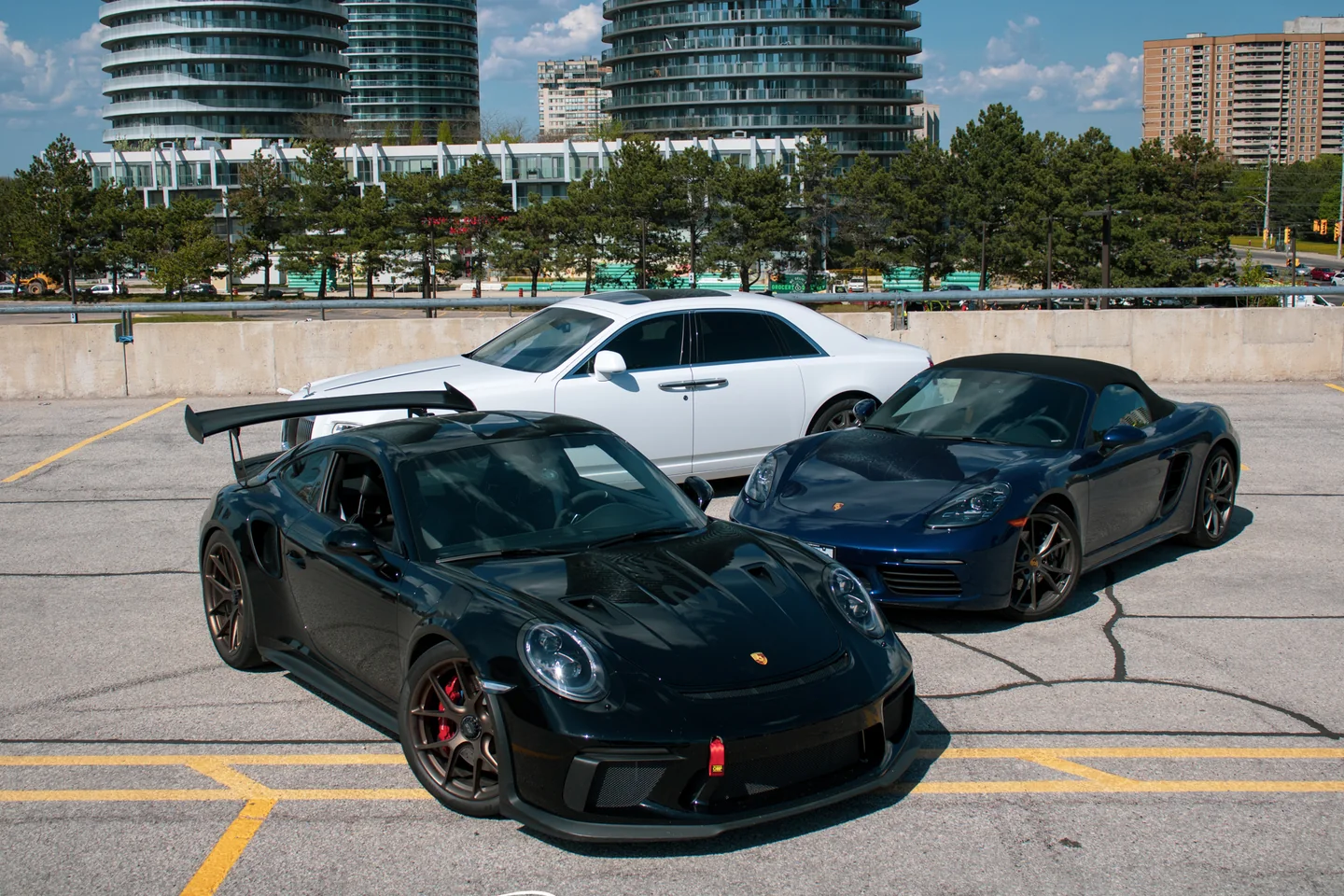 A black Porsche GT3 RS, a white car and a blue Boxster parked across an open deck above the city.