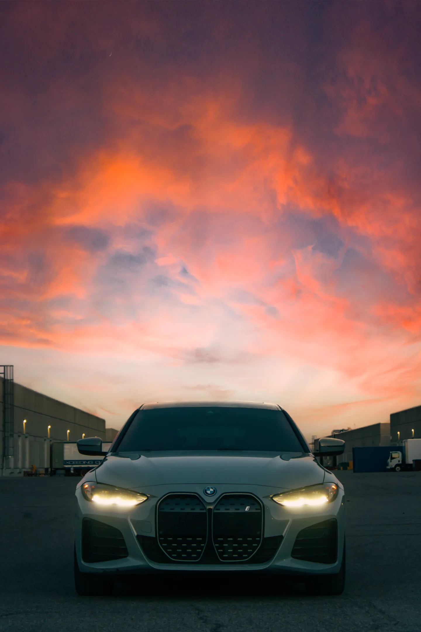 A BMW i4 head-on with its headlights lit under a sky burning pink and orange.