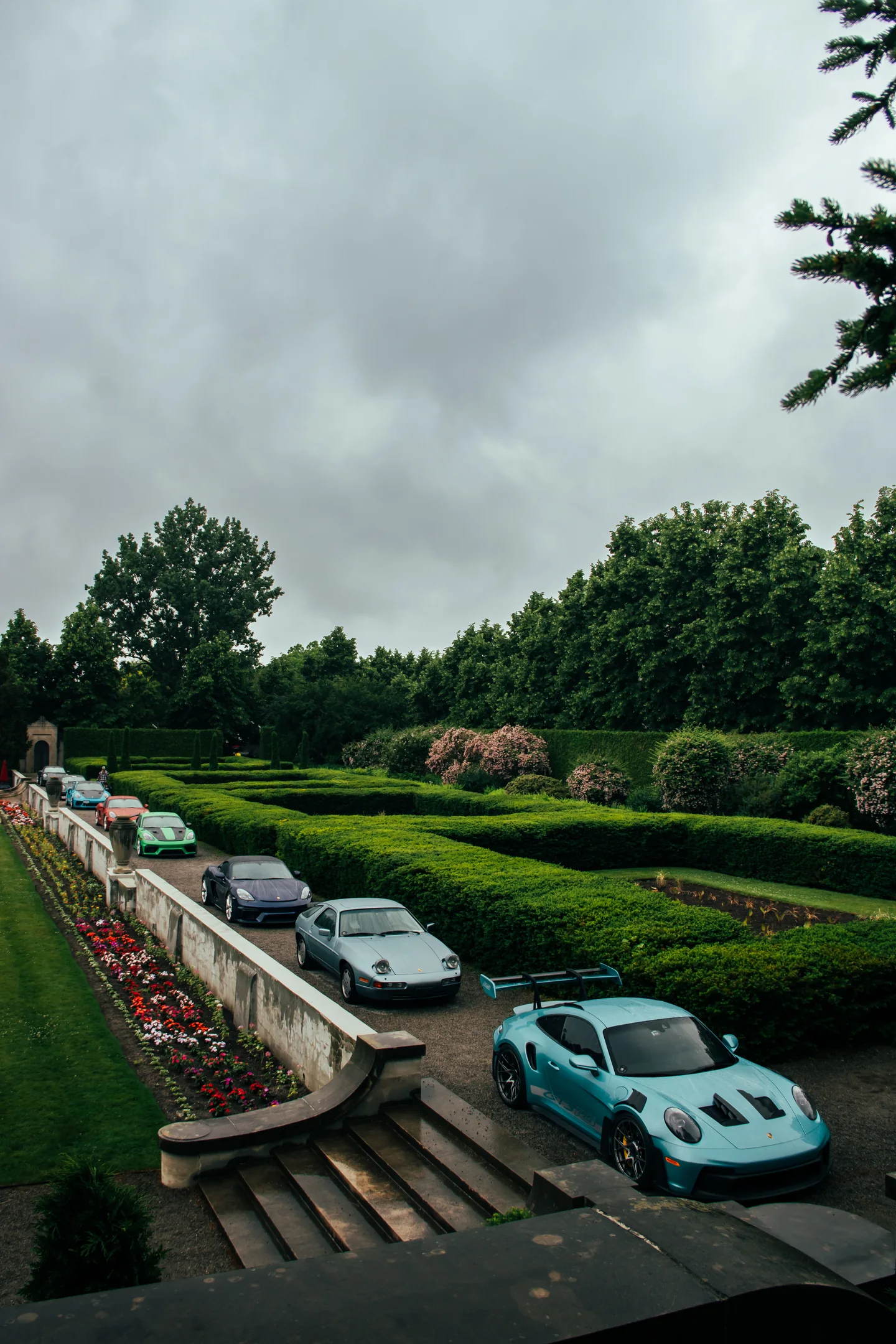 Cars parked along the formal garden terraces of a stone estate on an overcast morning.