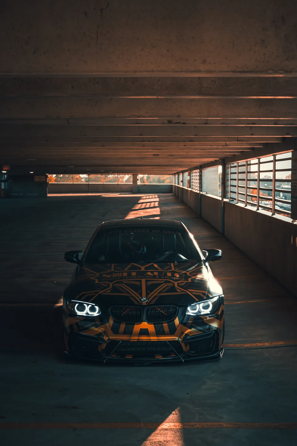 A liveried BMW on a parking deck at dusk, headlights on, concrete receding behind it.