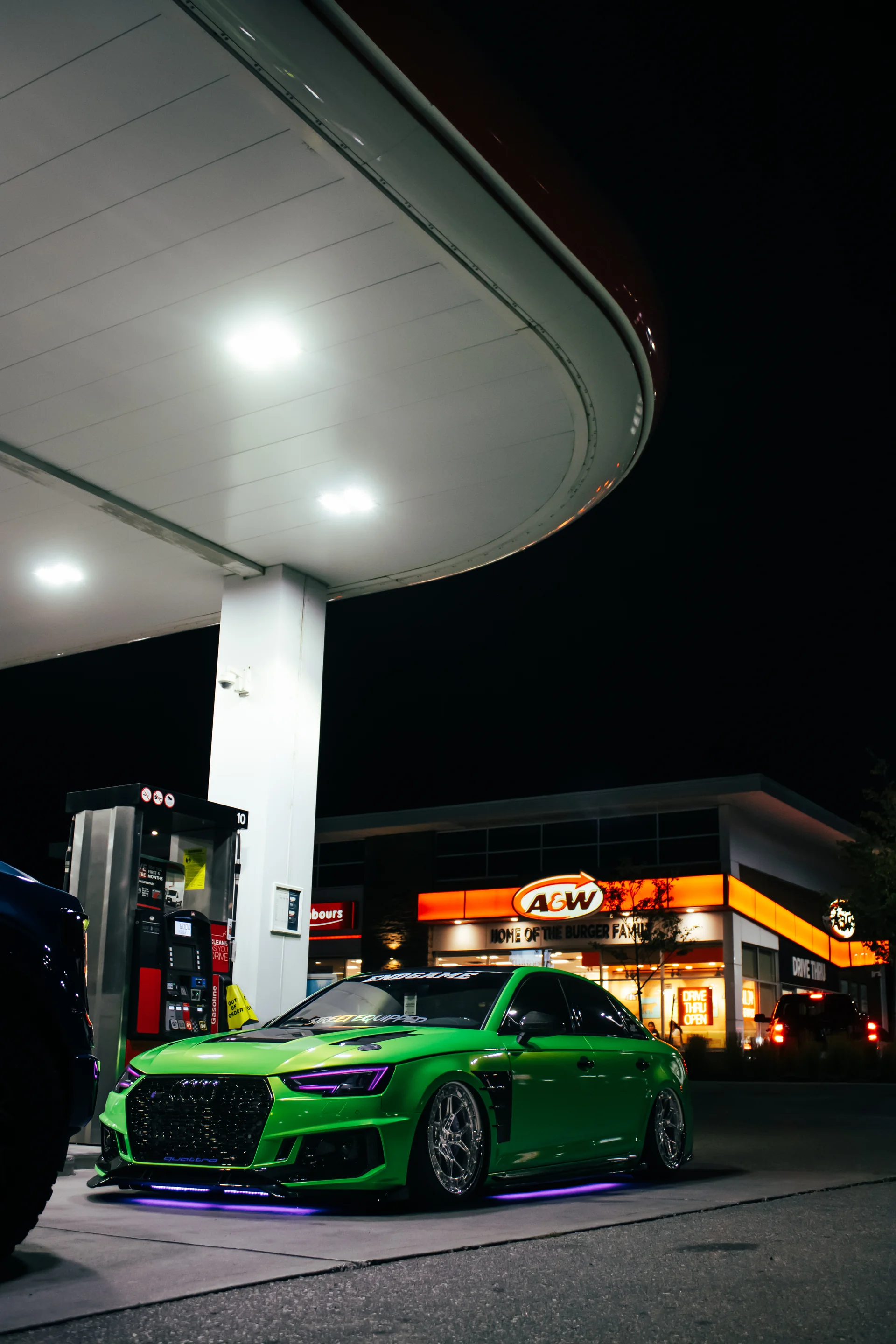 A green Audi S4 head-on beneath a gas station canopy with lit signage burning behind it.