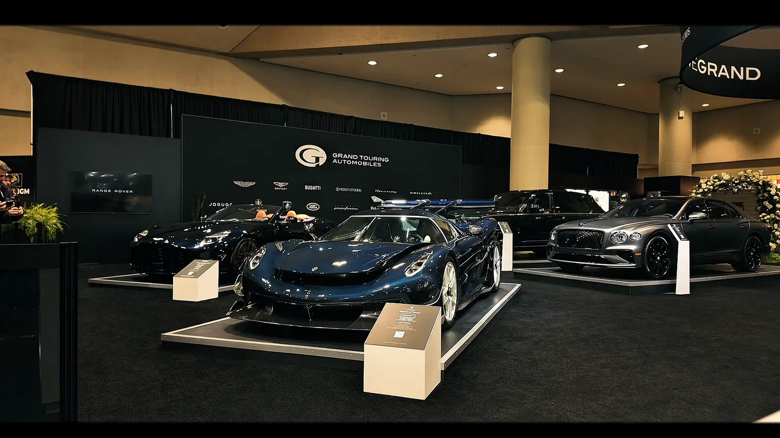 A dark blue hypercar raised on a show stand with its doors open, the camera moving slowly toward it across an auto show floor.