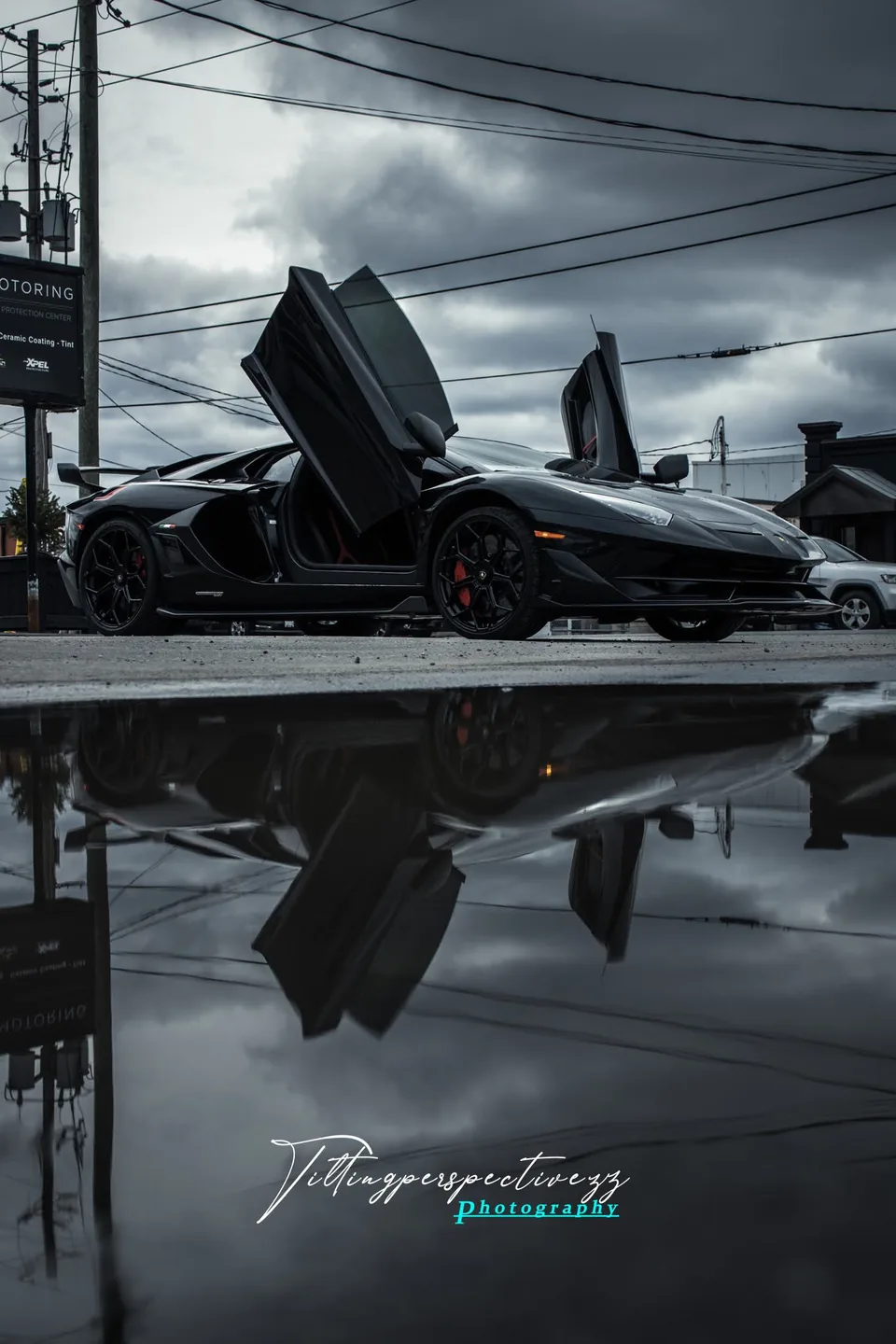 A Lamborghini SVJ with its doors raised, reflected in wet asphalt under a heavy sky.
