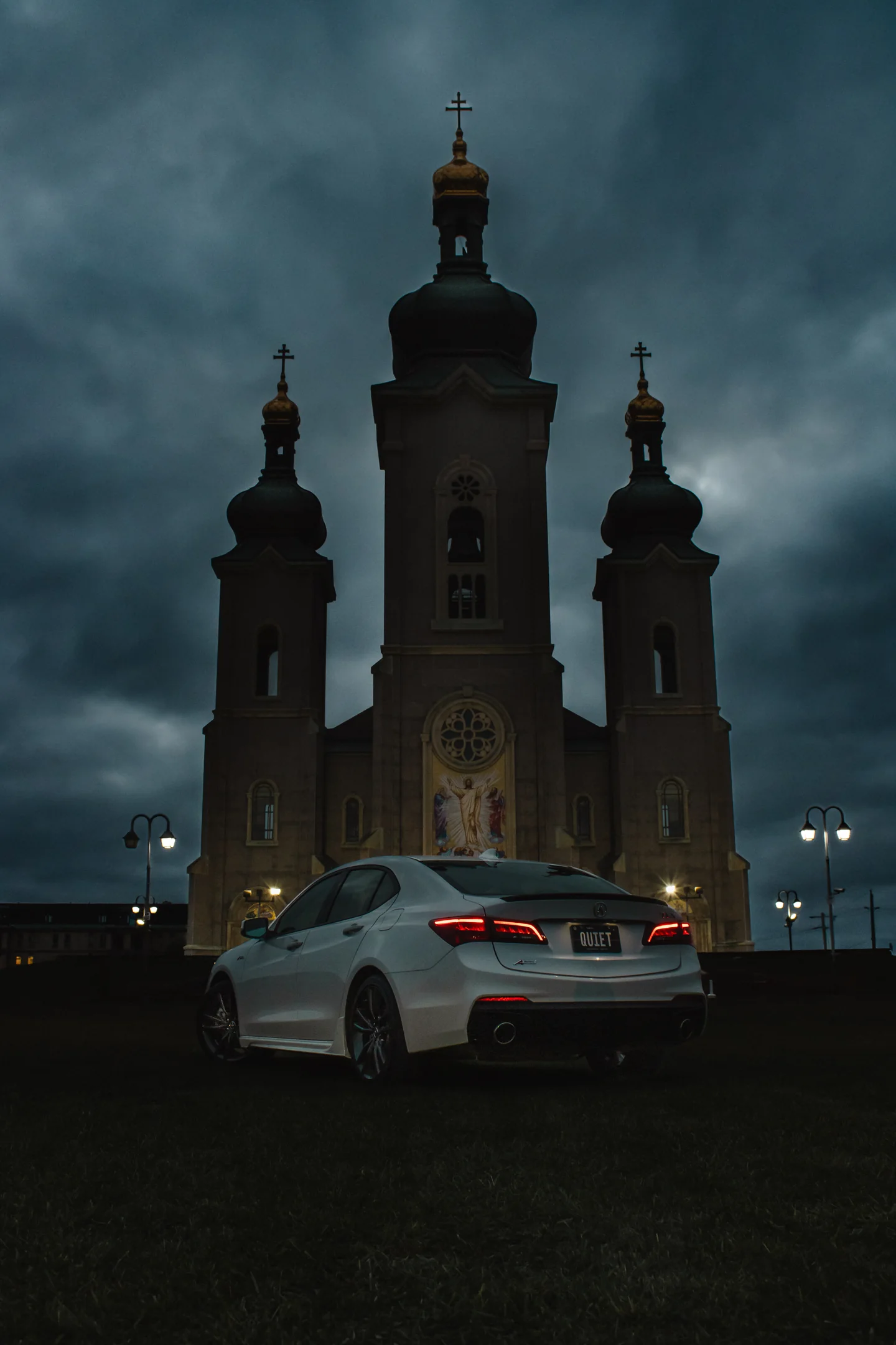 A white Acura TLX parked in front of a lit church at blue hour under moving cloud.