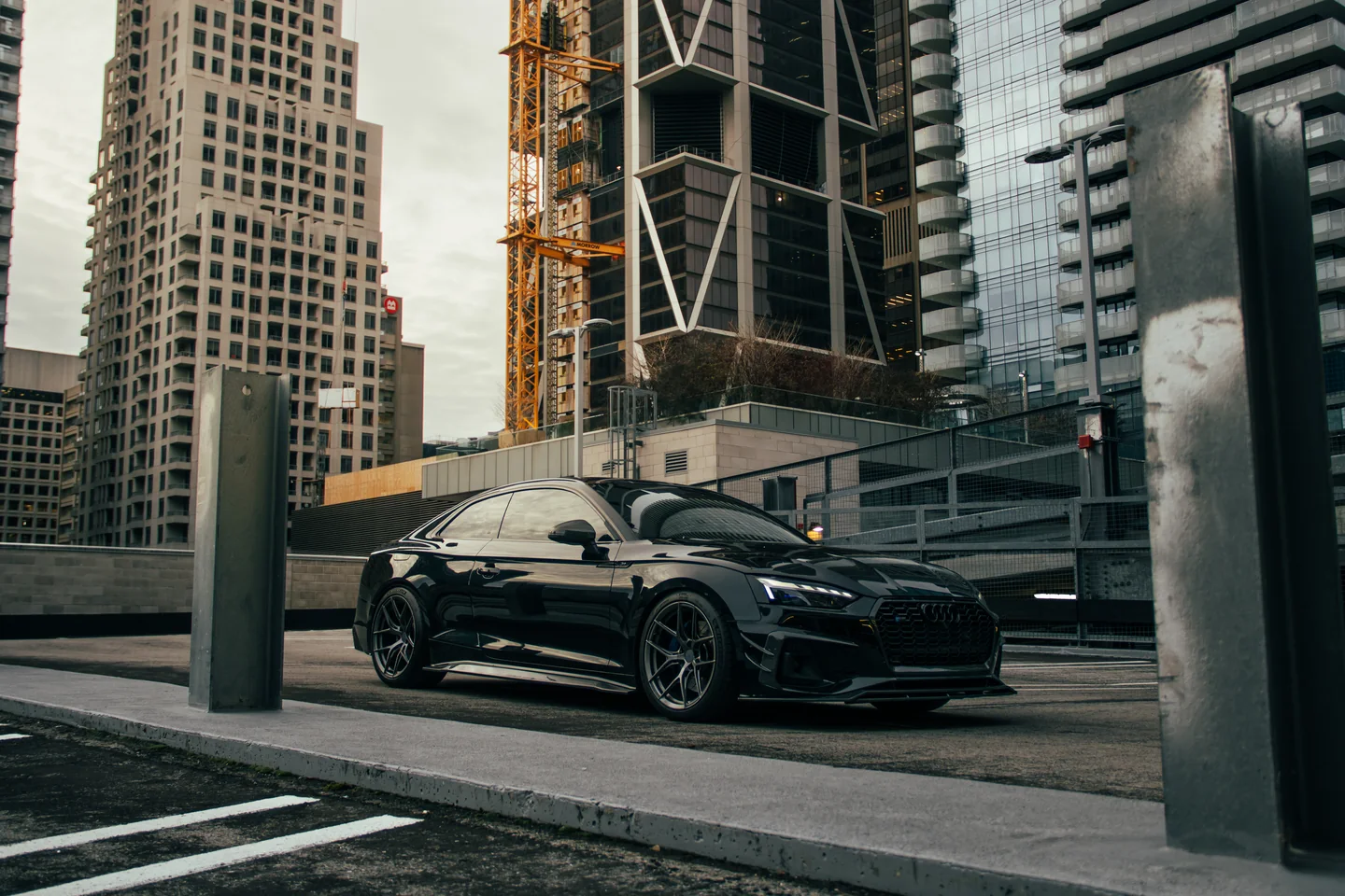 A black Audi S5 parked between concrete barriers with the Toronto skyline and a construction crane behind.