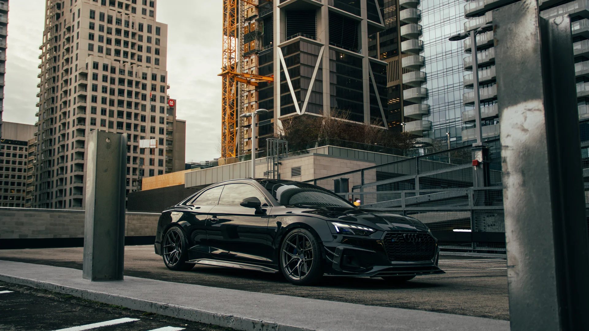 A black Audi S5 parked between concrete barriers with the Toronto skyline and a construction crane behind.