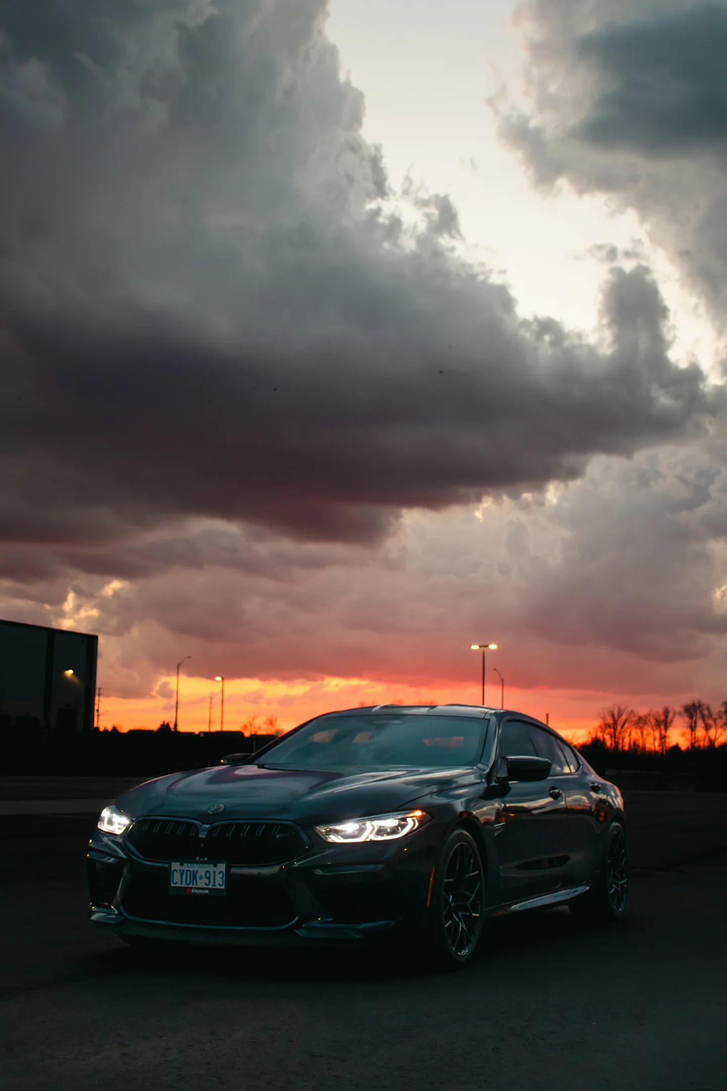 A BMW M8 Competition head-on at dusk with headlights lit beneath a burning storm front.