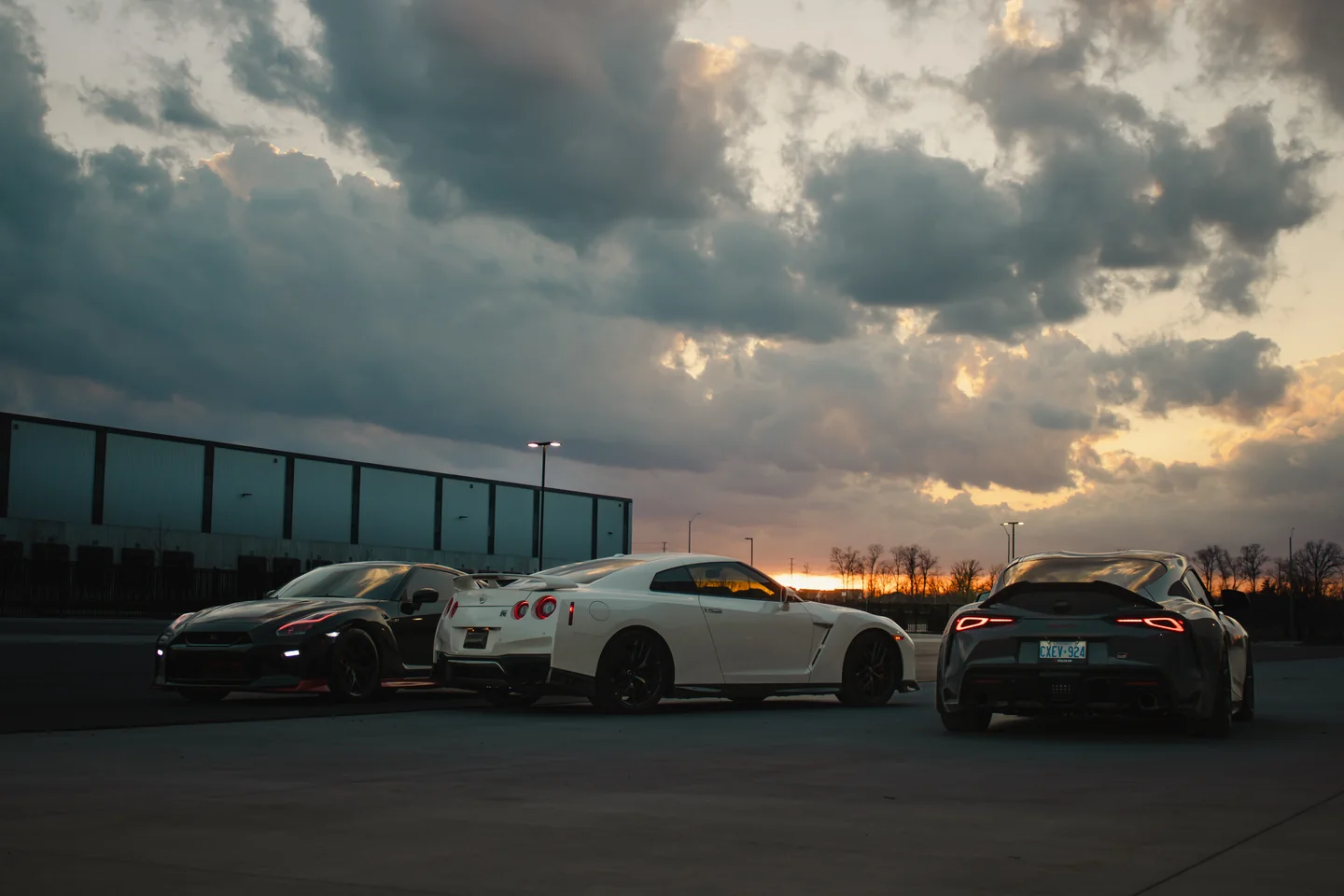 A GT-R, a second GT-R and a Supra lined up on open tarmac as the sun breaks under a heavy cloud bank.