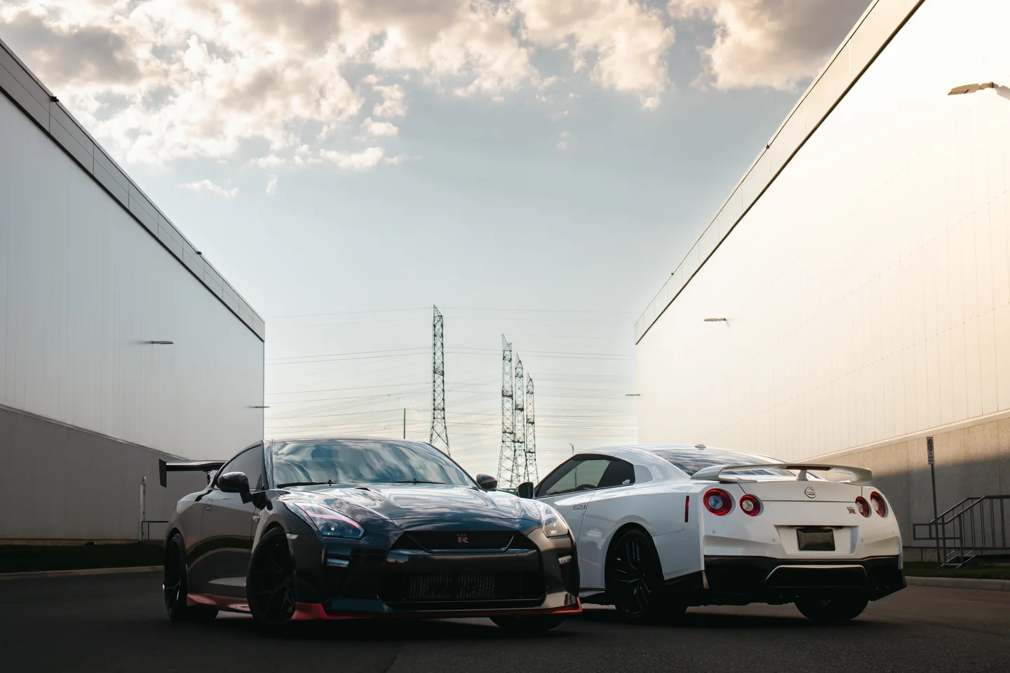 A black GT-R and a white GT-R parked between two pale industrial walls with hydro towers behind.