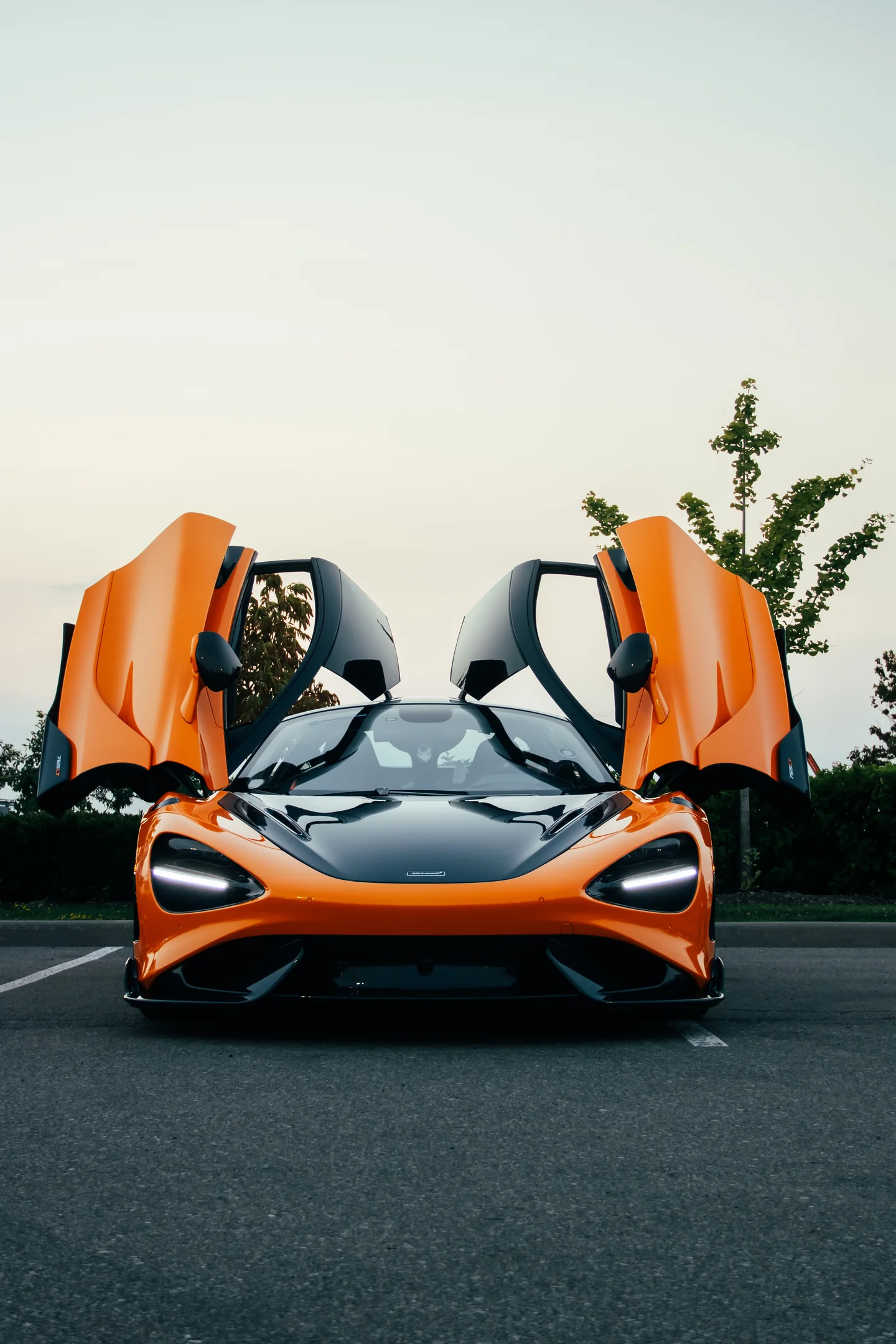 An orange McLaren head-on with both doors raised, trees behind it.