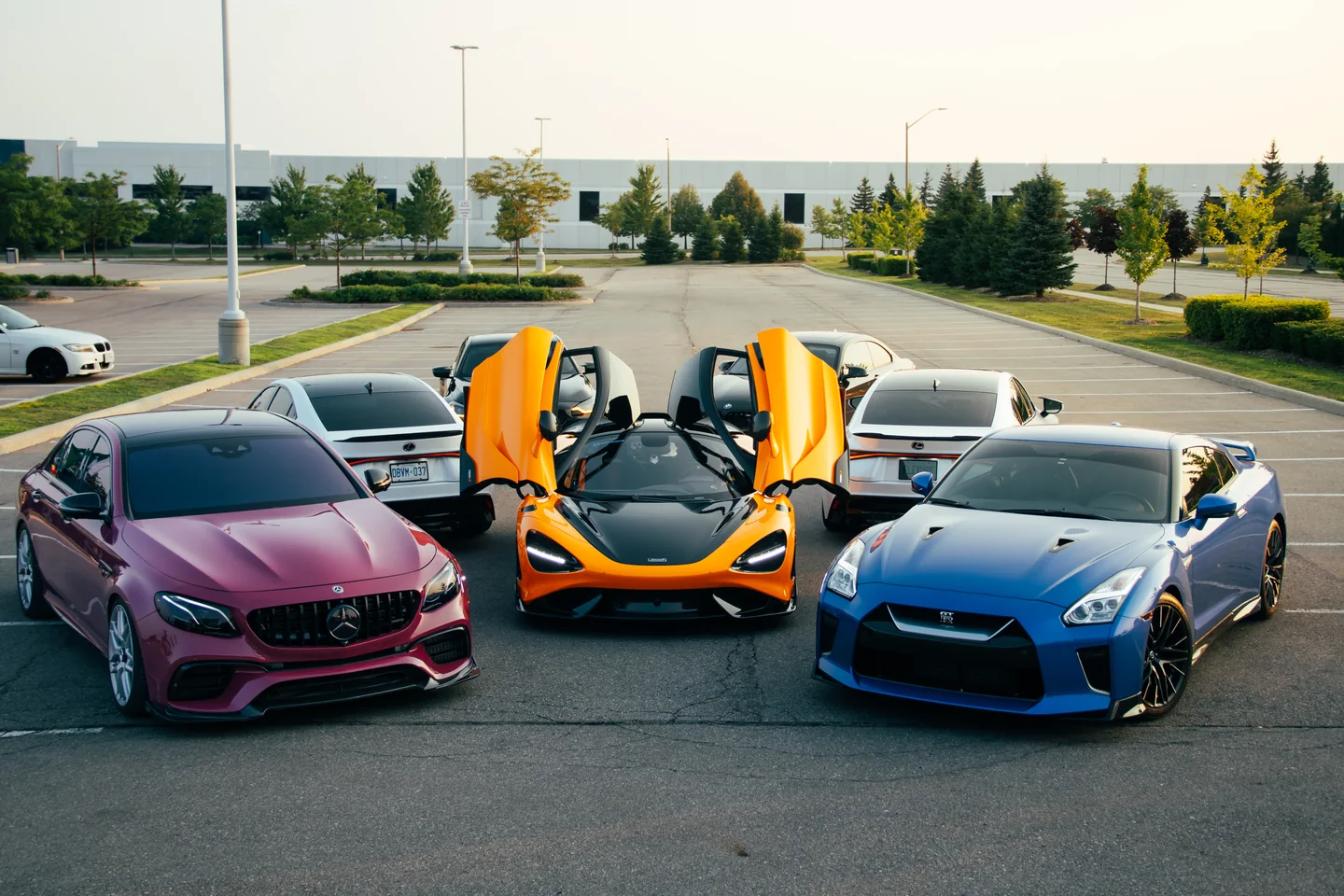 A group of cars parked with doors raised on an open lot by the water: AMG, McLaren, GT-R and more.