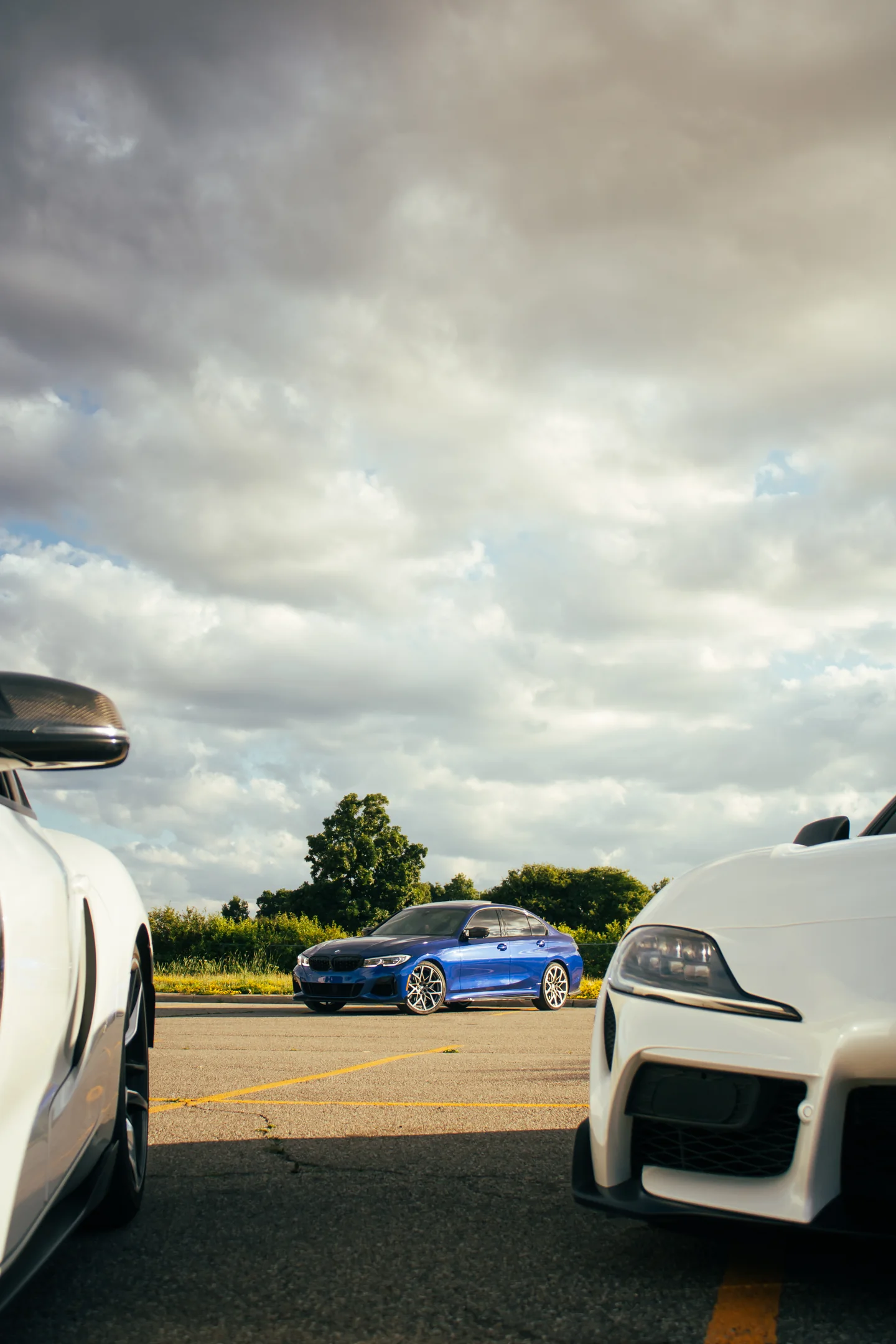 Two white Toyota Supras framing a blue BMW between their front ends under a broken sky.